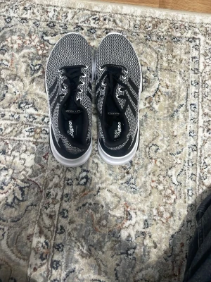 Svarta och vita sneakers från Adidas - Säljer ett par snygga Adidas sneakers i svart och vitt med stickad ovandel och klassiska tre ränder på sidan. Skorna har svarta snören och en vit sula som ger en sportig look. Perfekta för dig som gillar stilrena och bekväma sneakers.