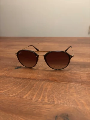 Ray-Ban solglasögon med bruna glas - Snygga Ray-Ban solglasögon med runda bruna glas och tunna guldfärgade metallbågar. Klassisk design med diskret logga på linsen och smala skalmar. Perfekta för att lyfta din stil under soliga dagar. Modell heter Rb 4292-n 710/13 61-14 Ordpris 2300kr Använda ett tiotal gånger