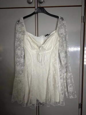 Vit spetsklänning med puffärmar - Romantisk vit klänning med transparenta spetsärmar och utsvängda ärmslut. Klänningen har figurnära passform, fyrkantig urringning och dekorativ knytning framtill. Tunt blommigt spetsmaterial över hela klänningen ger en drömmig look. Perfekt för dig som gillar vintagevibbar.