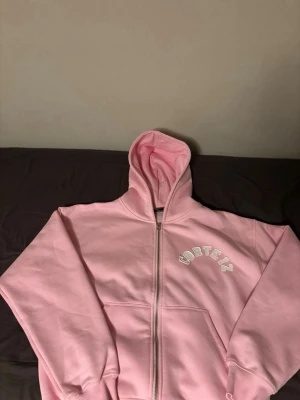 Rosa Corteiz zip hoodie med huva - Säljer en ljusrosa hoodie från Corteiz med dragkedja och stor huva. Hoodie har vita broderade Corteiz-loggor både fram och bak. Tillverkad i mjukt bomullsmaterial och har ribbade muddar vid ärmslut och nederkant. Perfekt för en chill och trendig look.