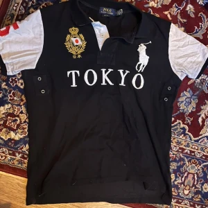 Polo Ralph Lauren Chief Keef Tokyo piké - Säljer en svart pikétröja från Polo Ralph Lauren med vita ärmar och stora broderade detaljer. Framsidan har TOKYO-tryck, guldigt emblem och den klassiska polospelaren. Baksidan har en stor röd femma. Tröjan har korta ärmar och knappar vid halsen.
