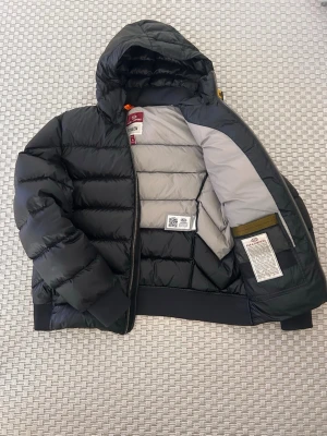 Svart dunjacka från Parajumpers - Snygg svart dunjacka från Parajumpers med huva och gul detalj vid halsen. Jackan har quiltad design, två sidofickor med dragkedja och ribbade muddar. Insidan är ljusgrå och jackan har klassisk Parajumpers-logga på ärmen. Perfekt för kalla vinterdagar.