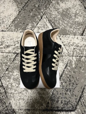 Svarta sneakers från maison margiela  - Stilrena svarta sneakers med kontrasterande vita snören och klassisk brun gummisula. Ovandelen är i en mix av mocka och slätt läder, vilket ger en snygg retrovibe. Perfekta för dig som gillar minimalistisk och tidlös design. Priset kan diskuteras!