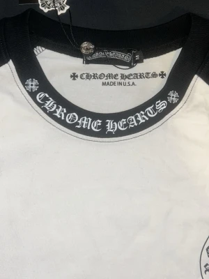Chrome Hearts svartvit långärmad tröja - Säljer en svartvit långärmad tröja från Chrome Hearts med tydlig logga tryckt på kragen och bröstet. Tröjan har svarta ärmar och krage, samt vit framsida. Tillverkad i bomull och har en cool streetwear-vibe.