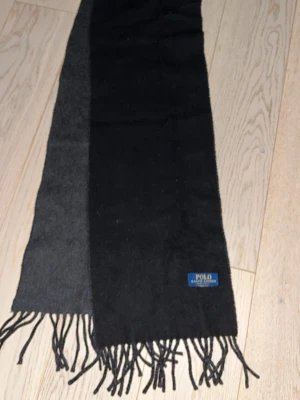 Svart och grå halsduk Polo Ralph Lauren - Snygg halsduk från Polo Ralph Lauren i svart och mörkgrå färg. Halsduken har klassiska fransar i ändarna och en diskret logga. Perfekt accessoar för att lyfta din outfit under kyliga dagar.