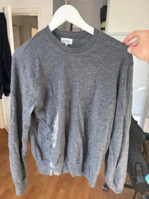 Grå stickad tröja från SANDÖ - Mysig grå stickad tröja från SANDÖ med rund halsringning och långa ärmar. Tröjan har en enkel och stilren design med ribbade muddar vid ärmslut och nederkant. Perfekt för lager på lager och passar till det mesta i garderoben.p