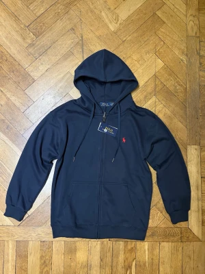 Mörkblå hoodie från Polo Ralph Lauren - Snygg mörkblå hoodie från Polo Ralph Lauren med dragkedja framtill och klassisk röd logga broderad på bröstet. Hoodien har huva med dragsko, två fickor fram och ribbade muddar vid ärmar och nederkant. Perfekt för en avslappnad och stilren look.
