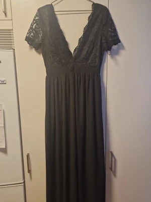 Svart långklänning med spetsdetaljer - Elegant svart långklänning med djup v-ringning och spetsdetaljer upptill. Klänningen har korta ärmar, markerad midja och en lång, flytande kjol. Dragkedja i ryggen för enkel påklädning. Perfekt för speciella tillfällen.