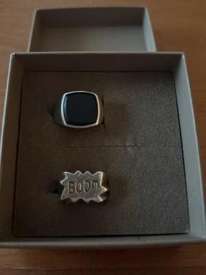 Två coola ringar i silverfärg - Säljer två unika ringar i silverfärgad metall. Den ena har en svart, fyrkantig sten och den andra har en serietidningsinspirerad design med texten 'BOOM'. Perfekt för dig som vill sticka ut med din stil. Strl M dvs 16,5-17
