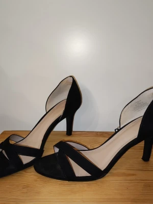 Svarta pumps med öppen tå från H&M - Stilrena svarta pumps från H&M med öppen tå och smala remmar i mockaimitation. Klackarna är medelhöga och skon har en elegant, klassisk form. Insidan är ljusbeige och ger en snygg kontrast till det svarta yttermaterialet.