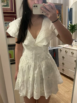 Studentklänning vit - Supersöt vit kort klänning med djup v-ringning och stora volangdetaljer vid axlarna. Klänningen har broderade blommor över hela tyget och markerad midja för en feminin siluett. Helt ny med prislapp