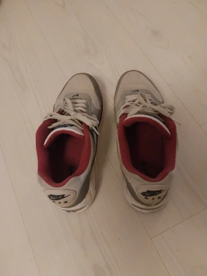 Nike Air Max 90 grå och röda sneakers - Nike Air Max 90 sneakers i grått, vitt och svart med röda detaljer och röd insida. Klassisk chunky sula med synlig Air Max-dämpning. Ovandel i mesh och läder, snörning framtill och Nike-logga på hälen. Inget synligt slitage. Använda av en dam. 