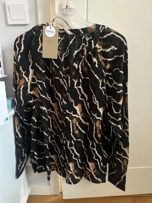 Mönstrad blus från Soyaconcept stl L - Snygg blus från Soyaconcept med svart bas och abstrakt mönster i brunt, beige och vitt. Blusen har en avslappnad passform, V-ringning och långa ärmar. Tillverkad i ett lätt och luftigt material med EcoVero-märkning.