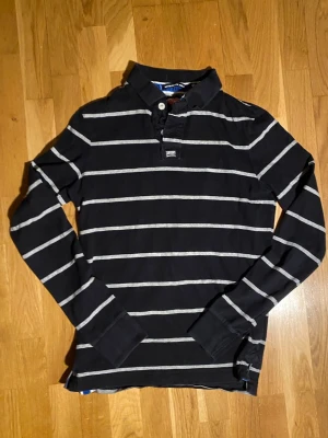 Superdry Randig Långärmad polotröja  - Snygg svart långärmad tröja med gråa horisontella ränder och krage. Tröjan har knäppning vid halsen. Den är från superdry. Längd 61 cm och bredd 45 cm. Ifall fler frågor eller bilder skriv!