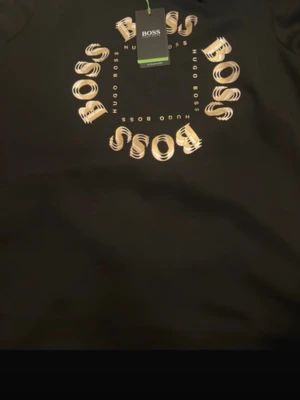 Svart t-shirt från Hugo Boss - Snygg svart t-shirt från Hugo Boss med coolt cirkelformat BOSS-tryck i beige och vitt på bröstet. Perfekt för dig som gillar stilrena plagg med en twist. Klassisk passform och mjukt material.