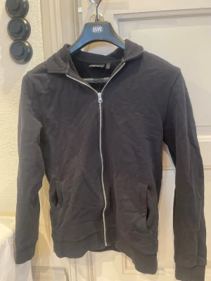 Svart ziptröja  - Svart ziptröja med hel dragkedja framtill och två fickor. Tröjan har en enkel krage och ribbade muddar vid ärmslut och nederkant. 