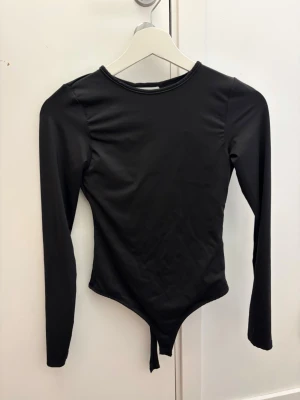 Svart body med öppen rygg - Säljer denna snygga långärmade bodyn från NewYorker som är använd 1 gång! Den är öppen i ryggen, har inbyggd kupor och har string ❣️ perfekt för en utekväll nu när det är kyligare! Passar också xs