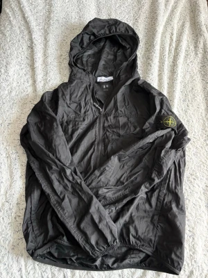 Svart vindjacka från Stone Island - Snygg svart vindjacka från Stone Island med klassisk patch på ärmen. Jackan har huva, sopren dragkedja framtill och elastiska muddar. Tillverkad i lätt och tåligt material som passar perfekt för blåsiga dagar.