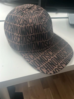 Brun keps med MOSCHINO-logga - Säljer en brun keps från Moschino med svart all-over logotyptryck. Klassisk form med böjd skärm och justerbar rem bak. Perfekt för dig som vill sticka ut med en trendig accessoar från ett välkänt märke.