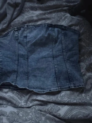 Blå denim bustier body med zip på ryggen - Säljer en snygg bustier body i blå denim med markerade sömmar och figurnära passform. Perfekt för dig som vill ha en trendig och edgy look. Materialet är klassisk jeansväv som ger en cool känsla. Inga synliga märken eller loggor.