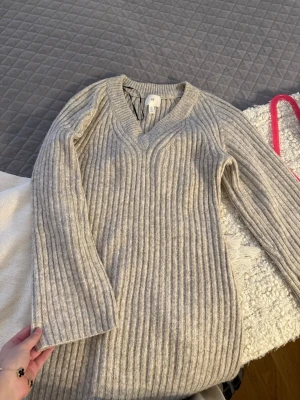 Beige ribbstickad klänning H&M - Lång, beige ribbstickad klänning från H&M med v-ringning och långa ärmar. Klänningen har en mjuk och stretchig känsla, perfekt för lager på lager. Enkel men snygg design som passar till många olika stilar.