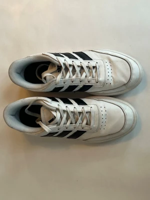 Vita Adidas sneakers med svarta ränder - Klassiska vita sneakers från Adidas med de ikoniska tre svarta ränderna på sidan. Skorna har lågt skaft, vita snören och ventilerande hål på tån. Tillverkade i läder och textil för en clean och sportig look.