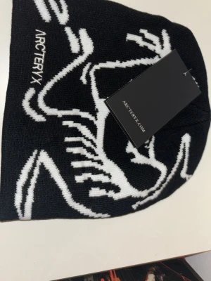Svart mössa från Arc'teryx - Säljer en svart mössa från Arc'teryx med vit grafisk design och logga. Mössan är stickad och har en clean, sportig look som passar perfekt till kalla dagar. Materialet är mjukt och bekvämt för huvudet.