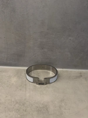 Silverfärgat armband med vit detalj - Stilrent armband i metall med silverfärgad finish och vita paneler på sidorna. Armbandet har en modern, minimalistisk design och ett smidigt spänne framtill. Perfekt accessoar för att lyfta din outfit med en clean look.