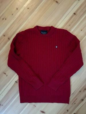 Röd kabelstickad tröja Bondelid - Röd kabelstickad tröja från Bondelid med rund halsringning och diskret logomärke på bröstet. Tröjan har långa ärmar och ribbade muddar vid ärmslut och nederkant. Perfekt för dig som gillar klassisk och stilren design.