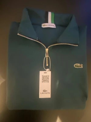 Mörkgrön zip hoodie från Lacoste - Snygg mörkgrön hoodie från Lacoste med halv dragkedja och klassisk krokodillogga på bröstet. Tröjan har ribbade muddar och krage med vit och grön detalj. Perfekt för en chill och sportig look. Materialet känns mjukt och bekvämt. Passar perfekt S