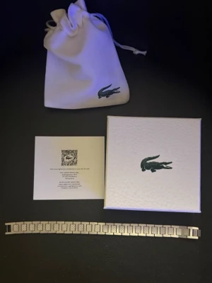 Silverfärgat armband från Lacoste - Snyggt armband i silverfärgad metall från Lacoste med stilrena, rektangulära länkar och graverad krokodil på spännet. Armbandet har en modern och clean design som passar perfekt till din outfit. Kommer med originalask och den är cirka 20cm lång kan fixa med en länkbortagare så den passar dig.