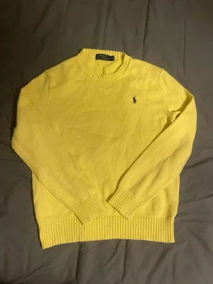 Gul stickad tröja från Polo Ralph Lauren - Gul stickad tröja från Polo Ralph Lauren med klassisk rund halsringning och den ikoniska mörkblå broderade loggan på bröstet. Tröjan har ribbade muddar vid ärmslut och nederkant. Perfekt för dig som vill ha en färgglad och stilren look.