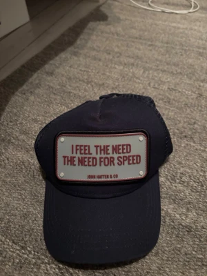 Mörkblå keps John Hatter & Co - Snygg mörkblå keps från John Hatter & Co med ett stort patch framtill där det står 'I FEEL THE NEED THE NEED FOR SPEED' i rött. Klassisk böjd skärm och mesh baktill för extra ventilation. Perfekt för dig som gillar statement-plagg.