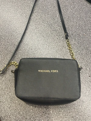 Svart axelväska Michael Kors - Snygg svart axelväska från Michael Kors i stilren boxig modell. Väskan har guldfärgade kedjedetaljer på axelremmen och logga i guld framtill. Tillverkad i tåligt konstläder med slät finish. Perfekt för dig som gillar klassisk och trendig design.