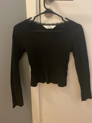 Svart långärmad tröja. Spetskrage. 10-12 år andvänd ca 3 gånger🌟 - Svart långärmad topp från H&M med ribbad struktur, spetskant vid urringningen och en liten rosett framtill. Figurnära passform och mjukt material som känns skönt mot huden. Perfekt för en stilren och trendig look.