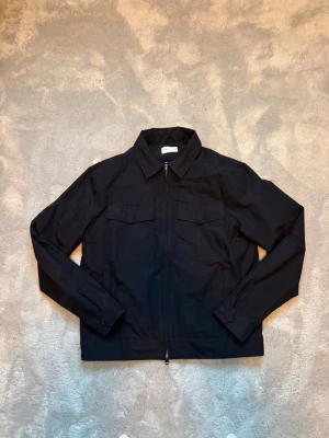 Svart overshirt med dragkedja från These Glory Days - Stilren svart overshirt med rak passform och klassisk krage. Två bröstfickor med lock framtill och hel dragkedja. Tillverkad i ett tunt, lätt material som passar perfekt för lager-på-lager. Långa ärmar med knapp vid ärmslut och minimalistisk design.