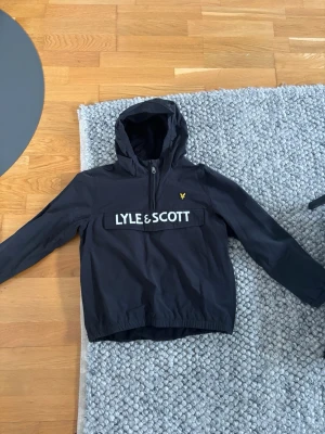 Svart vindjacka från Lyle & Scott - Snygg svart vindjacka från Lyle & Scott med huva och dragkedja framtill. Stor logga på bröstet och den klassiska gula örnen på vänster sida. Jackan har elastisk nederkant och är tillverkad i ett lätt material som passar perfekt till blåsiga dagar.