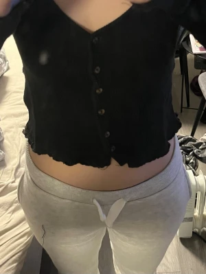 Svart croppad ribbad topp - Säljer en svart croppad topp med v-ringning och knappar framtill. Toppen är ribbad och har en vågig kant nertill. Perfekt för dig som gillar en enkel men snygg stil.