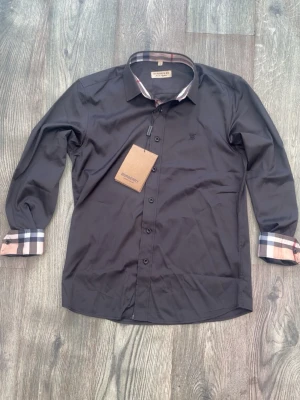 Svart skjorta från Burberry - Stilren svart skjorta från Burberry med klassiska rutiga detaljer på insidan av kragen, knappslån och manschetterna. Skjortan har lång ärm, knappar framtill och en diskret Burberry-logga broderad på bröstet. Perfekt för dig som gillar exklusiv och tidlös design.