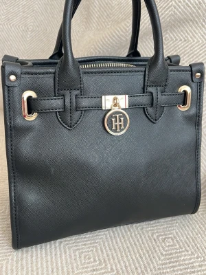 Svart handväska från Tommy Hilfiger - Stilren svart handväska i strukturerat konstläder från Tommy Hilfiger. Väskan har guldfärgade metalldetaljer, dubbla handtag och ett TH-logomärke framtill. Klassisk fyrkantig form och dragkedja upptill. Perfekt för dig som gillar tidlös design. Nypris är ca 2500kr