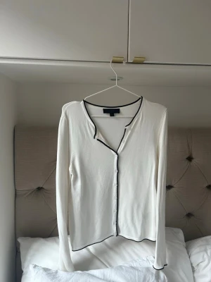 Vit kofta med svarta kanter - Snygg vit kofta med svarta kontrastkanter och knäppning framtill. Långärmad och med en smal passform, perfekt att styla till jeans eller kjol. Enkel men trendig design som passar till många olika looks.