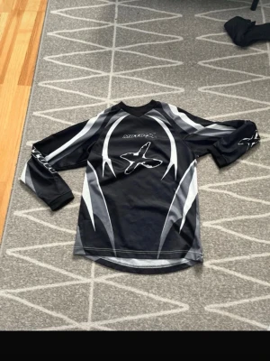 Svart Moto-X långärmad tröja - Säljer en svart långärmad tröja från Moto-X med grå och vita grafiska detaljer och stort X-tryck på bröstet. Tröjan har V-ringad hals och är tillverkad i ett lätt och ventilerande material, perfekt för motocross eller annan sport.