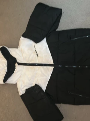 Svartvit pufferjacka från Only - Snygg svartvit pufferjacka från Only med huva och dragkedja framtill. Jackan har en cool färgblock-design, stora fickor med dragkedja och är vadderad för extra värme. Perfekt för kalla vinterdagar och ger en trendig look.