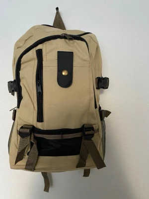 Beige ryggsäck med svarta detaljer - Snygg beige ryggsäck med flera fack och svarta dragkedjor. Framsidan har en dekorativ svart rem med guldfärgad knapp. Justerbara bruna remmar och meshfickor på sidorna. Perfekt för skolan eller utflykten.