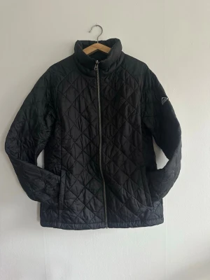 Svart quiltad vårjacka McKinley st 40 - Snygg svart  quiltat mönster och hög krage. Jackan har dragkedja framtill och två sidofickor. Perfekt för kalla dagar och ger en stilren look. Materialet är lätt och värmande, och jackan har en diskret logga på ärmen. Märkt med st 42 men motsvarar 40 bättre 