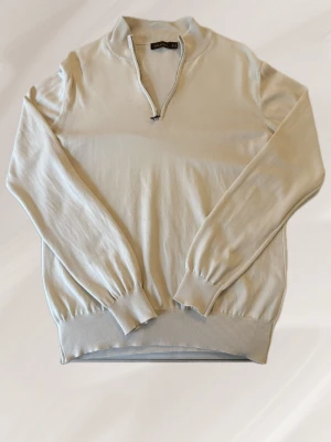 Beige quarterzip från oscar jacobson - Snygg beige stickad quarter-zip tröja från Oscar Jacobson. Tröjan har en hög krage. Perfekt för lager på lager och enkel att matcha till olika outfits. Passar kring M/S