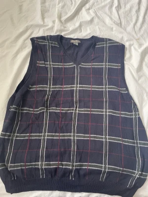 Mörkblå rutig väst från Eddie Bauer - Snygg mörkblå stickad väst från Eddie Bauer i 100% bomull. Västen har ett klassiskt rutmönster i vitt och rött samt v-ringad hals. Perfekt för lager-på-lager och ger en retrovibe till din outfit.