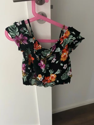 Blommig croppad offshoulder topp - Snygg croppad topp med offshoulder och smala axelband. Svart botten med färgglatt blommönster i lila, orange, grön och vit. Perfekt för sommaren och ger en fräsch vibe till din outfit.