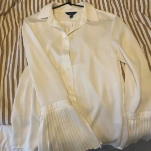 GANT Vit blus/skjorta med plisserade ärmar  - Stilren vit blus från KappAhl med klassisk krage och knäppning framtill. Blusen har långa ärmar med breda plisserade manschetter som ger en trendig touch. Tillverkad i ett mjukt, lätt material som faller snyggt.