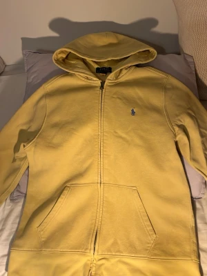 Gul hoodie från Polo Ralph Lauren - Gul hoodie med dragkedja från Polo Ralph Lauren. Klassisk blå logga på bröstet och stort blått Polo 67-tryck på ryggen. Två fickor fram och huva. Perfekt för en avslappnad och stilren look.